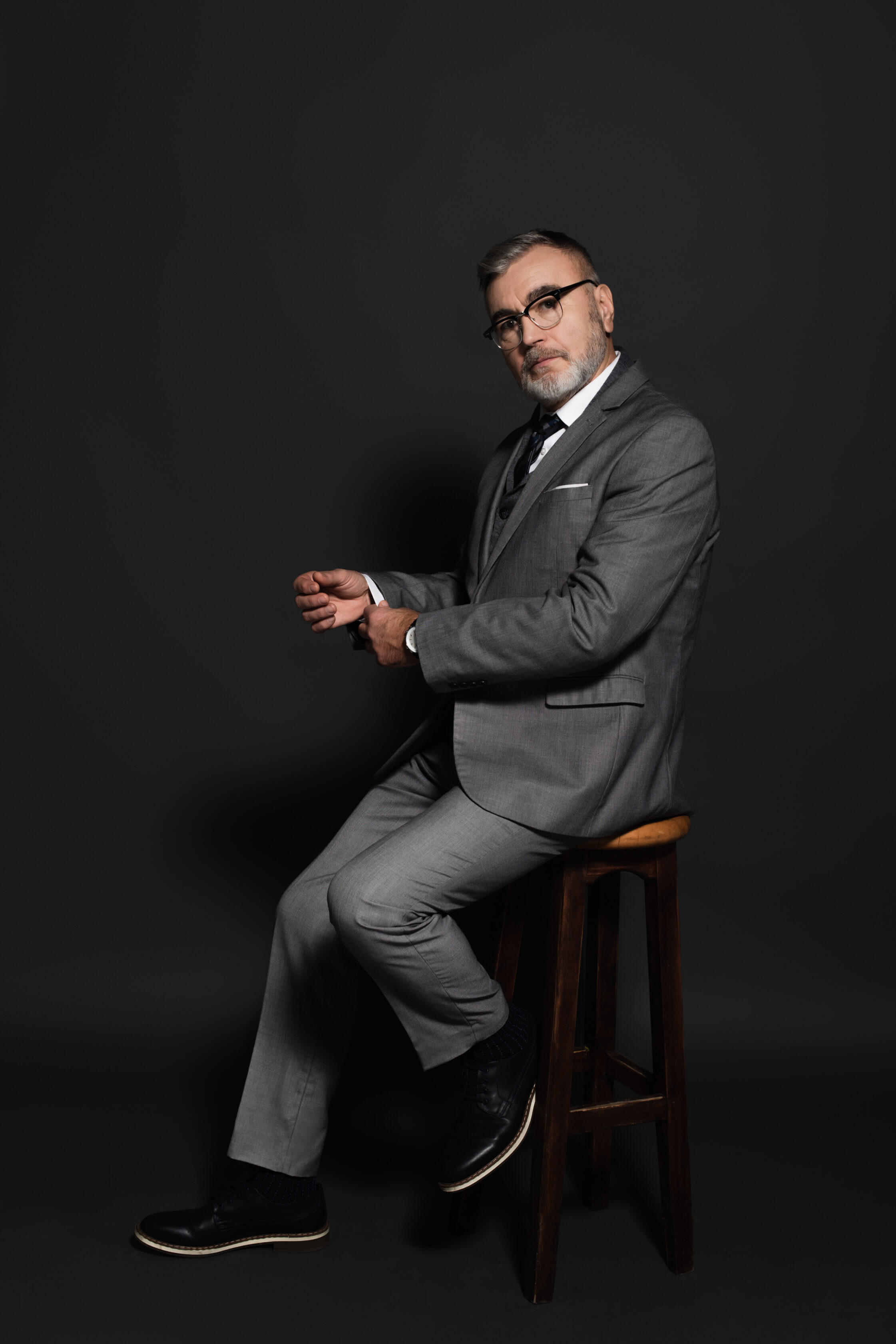 full length view of senior man in suit and eyeglasses sitting on high stool and looking at camera on